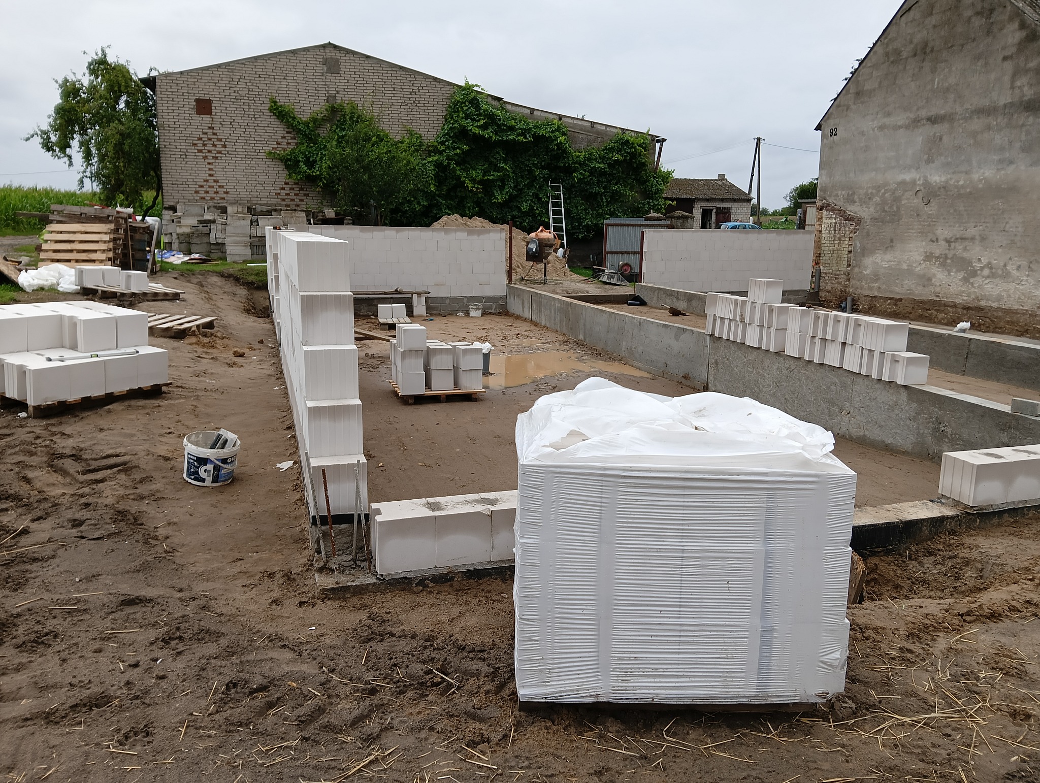 Budowa domu z betonu komórkowego: częściowo wymurowane ściany fundamentowe, paleta z bloczkami w folii, widoczny murarz przy pracy, budynek gospodarczy w tle.