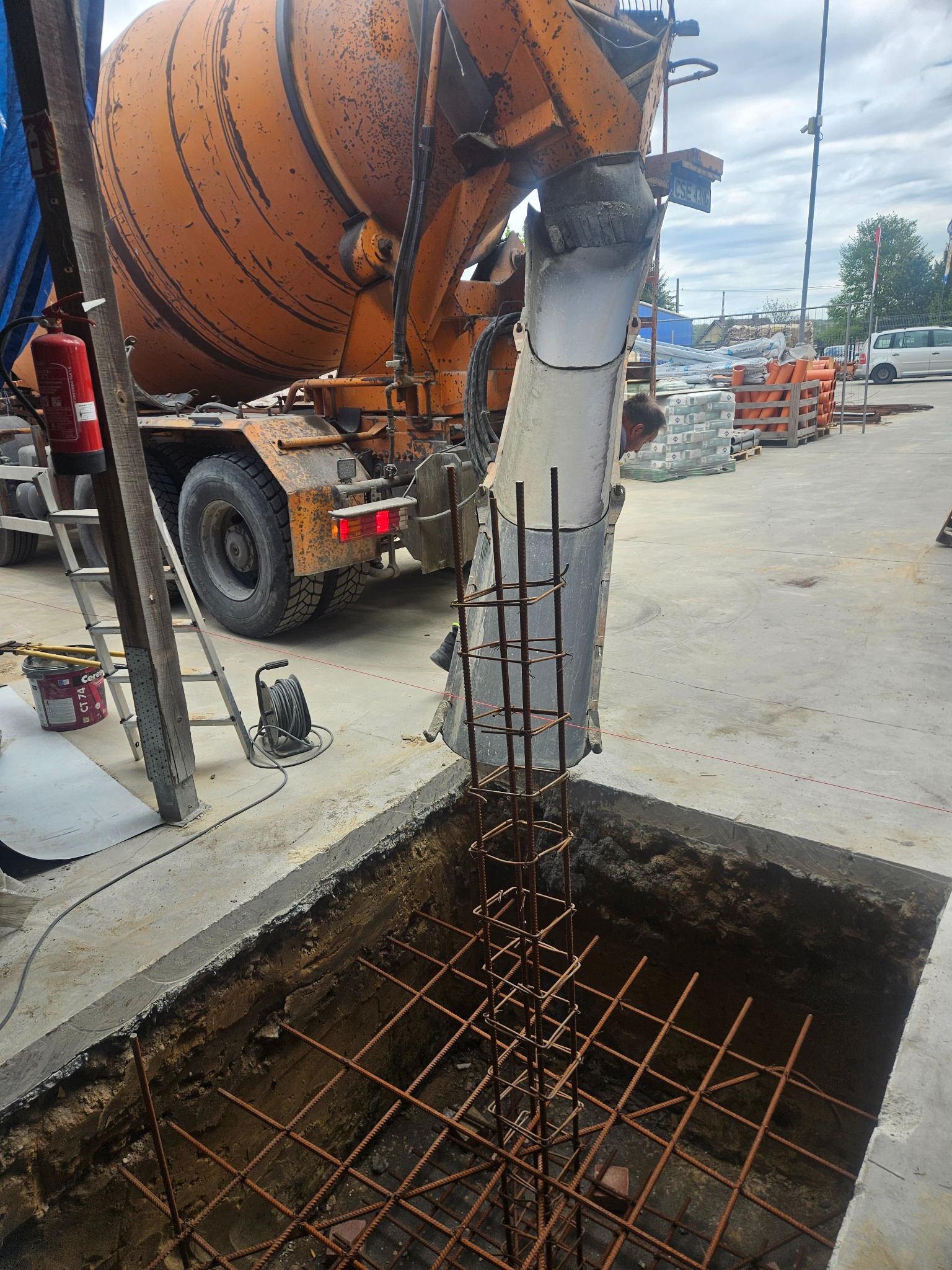 Betoniarka wylewa beton do wykopu zbrojonego stalą. Widoczna drabina, wiadro i zwinięty kabel. W tle budynki i błękitne niebo.