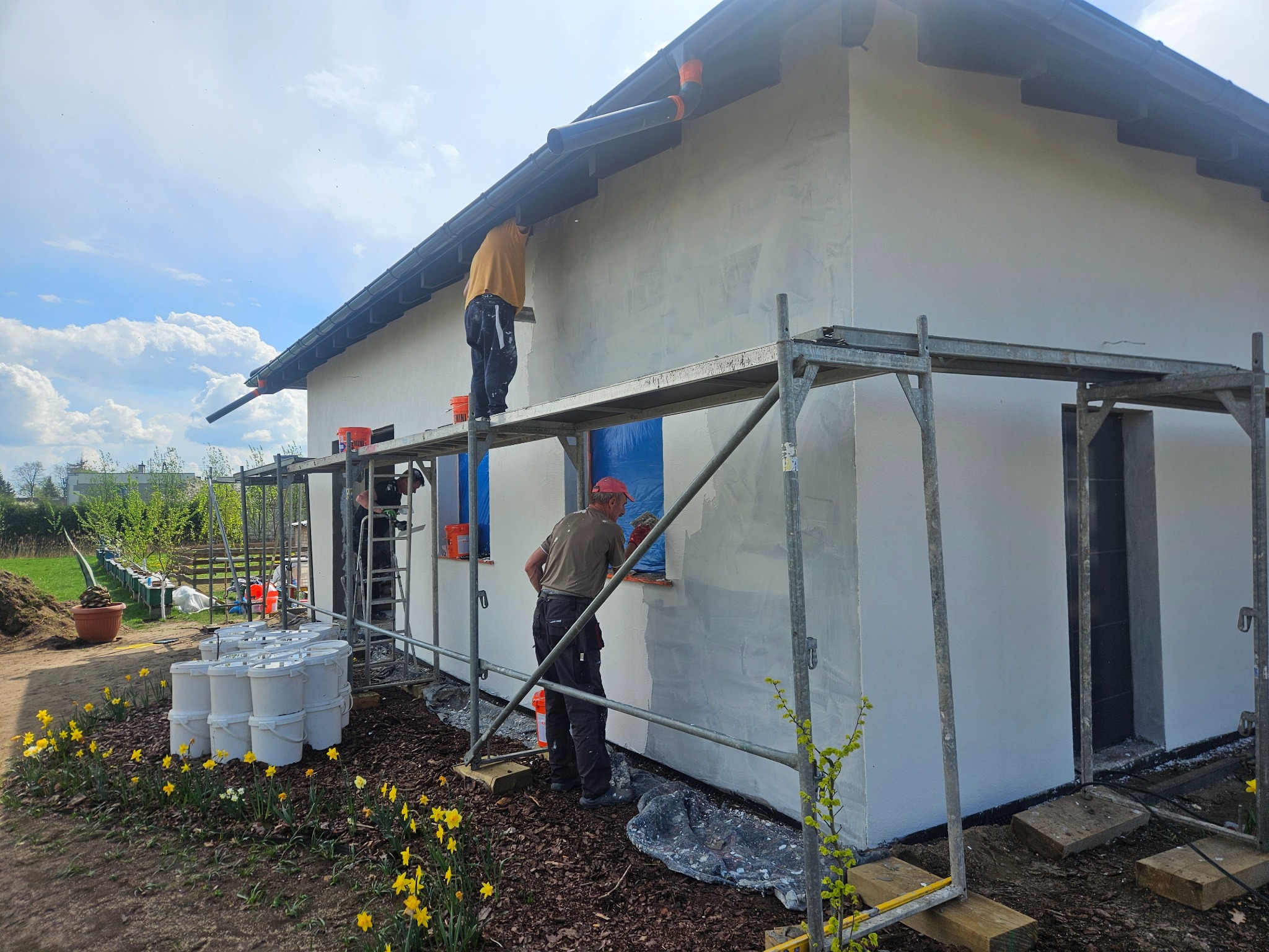 Dwóch pracowników na rusztowaniu tynkuje elewację domu jednorodzinnego w słoneczny dzień. Widoczne wiaderka z zaprawą i świeżo posadzone kwiaty.