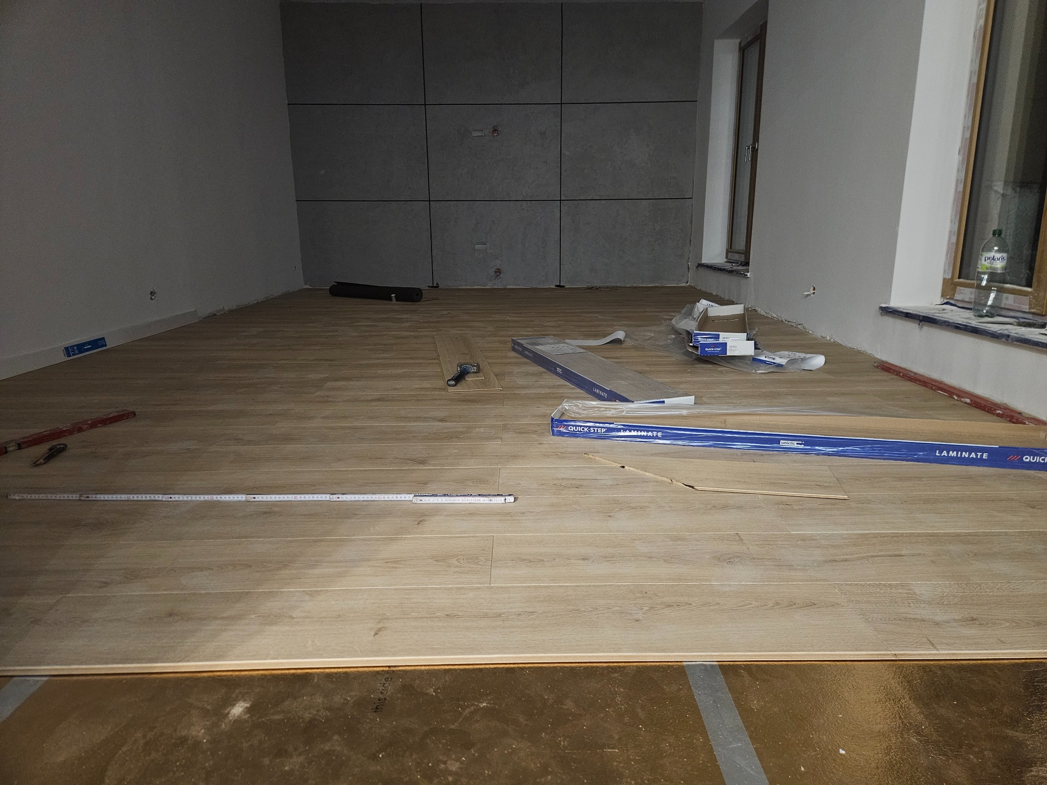 Układanie paneli podłogowych w jasnym pomieszczeniu z szarą ścianą akcentującą. Widoczne narzędzia i opakowania paneli Quick-Step. Częściowo ułożona podłoga na podkładzie.