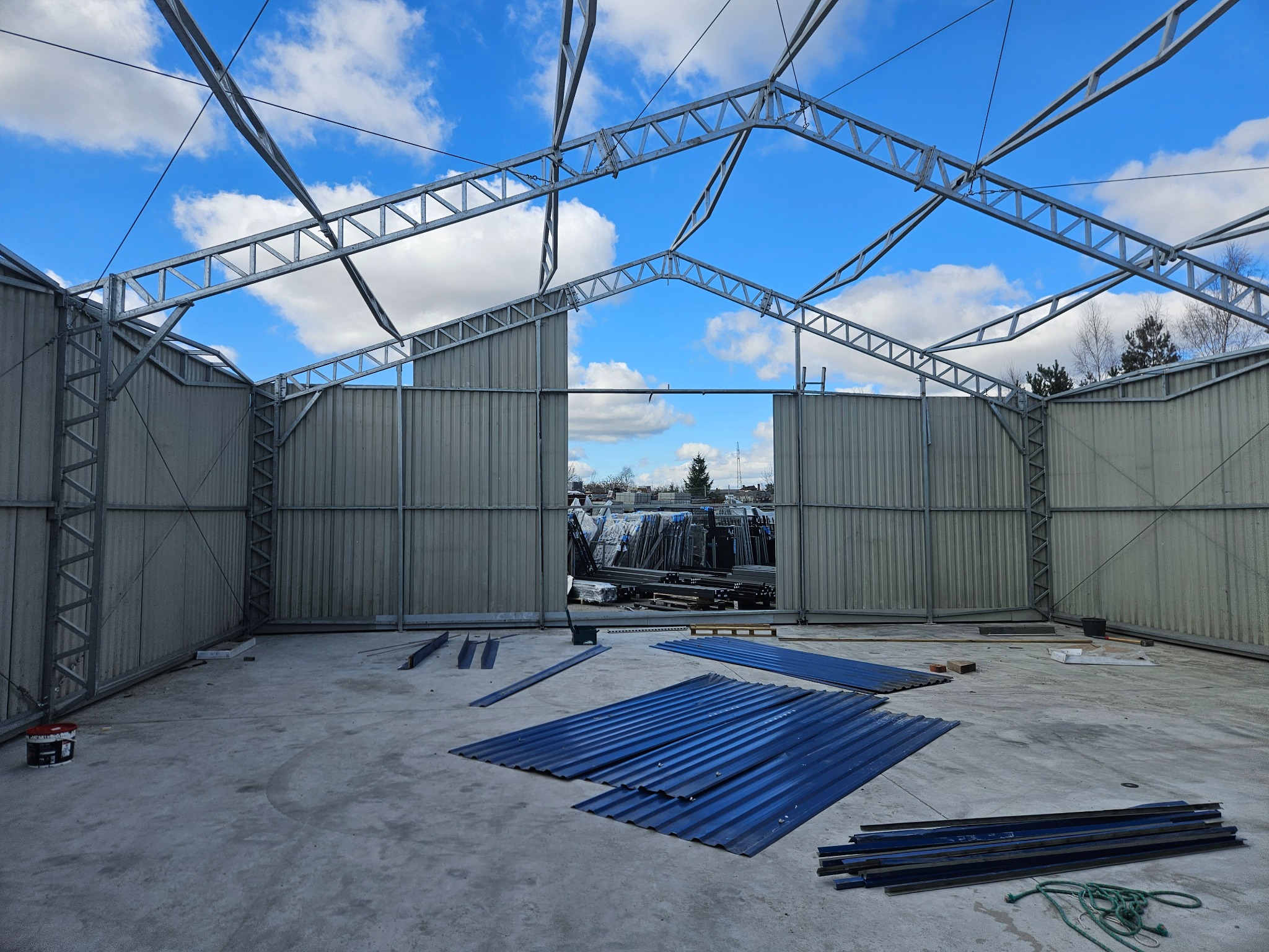 Konstrukcja stalowa hali w trakcie montażu, widoczne elementy poszycia i niebo w tle. Na posadzce leżą arkusze blachy trapezowej.