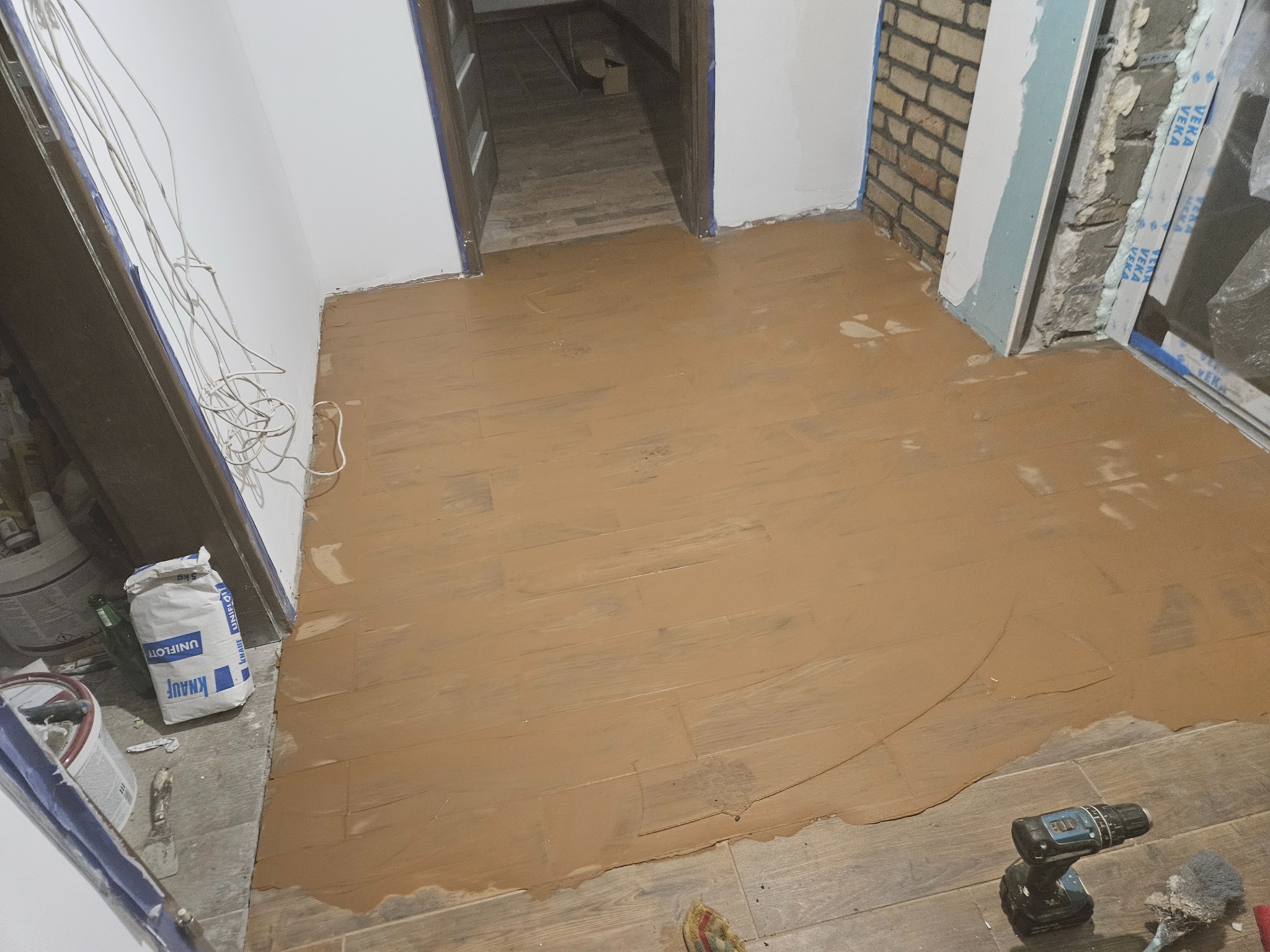 Wyrównywanie podłogi w korytarzu brązową masą szpachlową na drewnopodobnych płytkach. Widoczny worek z zaprawą, wiertarka i narzędzia.