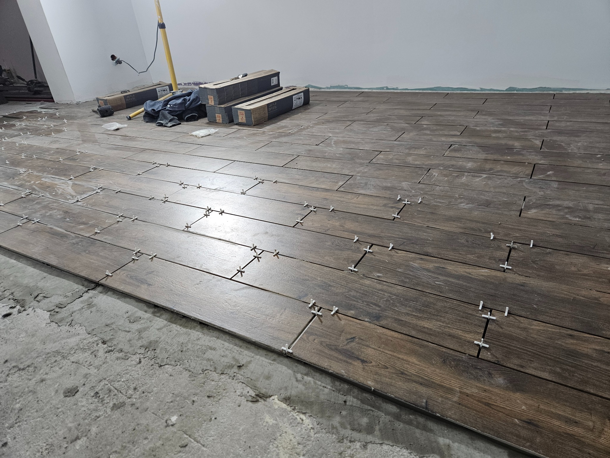 Układanie podłogi z płytek imitujących drewno, z użyciem krzyżyków dystansowych. Widoczne kartony z płytkami i poziomica. Częściowo ułożona podłoga na surowym podłożu.