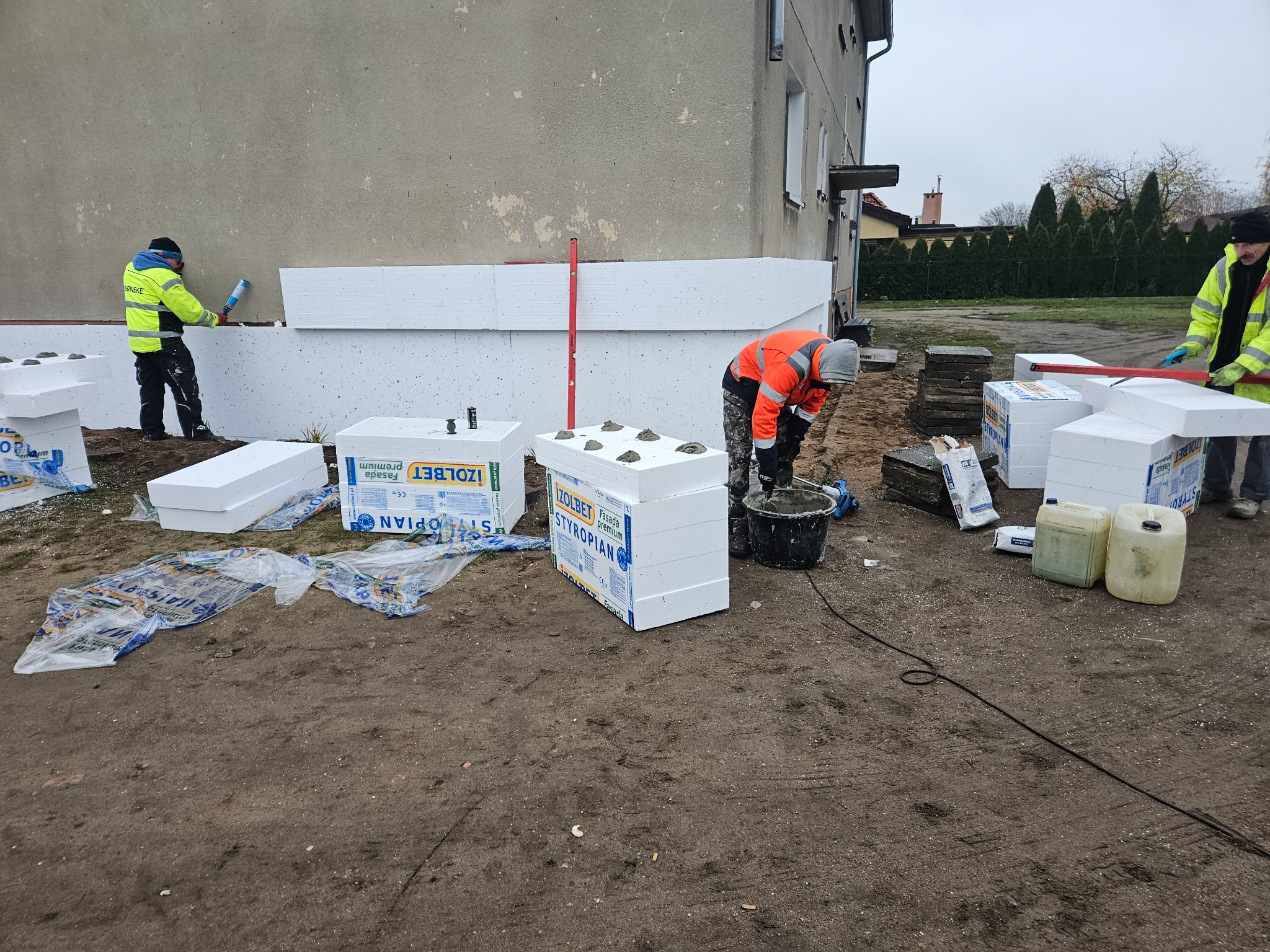 Ocieplanie fundamentów budynku styropianem IzoLbet. Dwóch pracowników w odblaskowych ubraniach przy pracy. Widoczne bloki styropianowe i narzędzia.