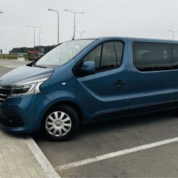 V-car rent - Niebieski bus Renault Trafic na parkingu z kostki brukowej, widok z boku, pochmurne niebo w tle. Widoczne światła uliczne i zieleń w oddali.