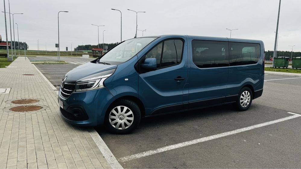 Niebieski bus Renault Trafic na parkingu z kostki brukowej, widok z boku, pochmurne niebo w tle. Widoczne światła uliczne i zieleń w oddali.