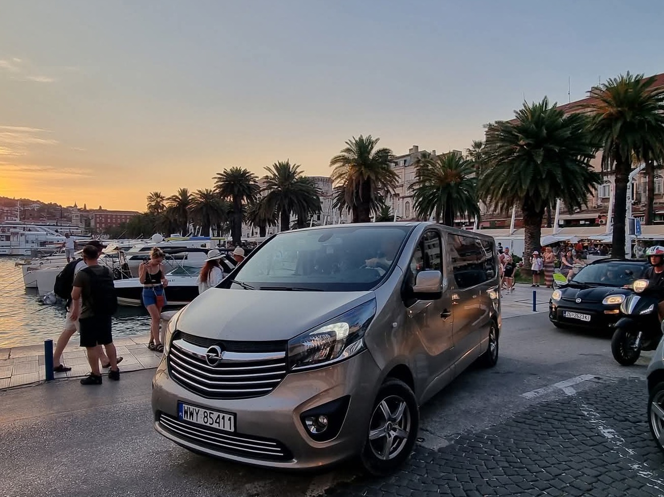Szary Opel Vivaro na tle portu jachtowego i palm w Chorwacji. W tle zachód słońca i spacerujący ludzie. Tablica rejestracyjna WWY 85411.