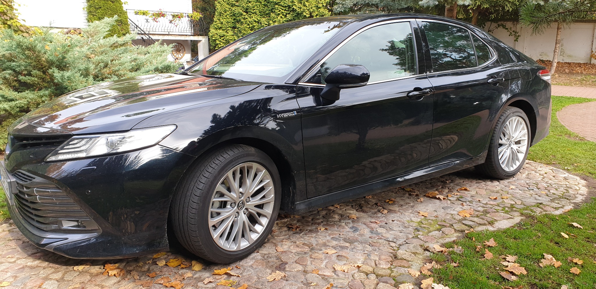 Czarna Toyota Camry Hybrid na brukowanej nawierzchni, jesienne liście, zieleń w tle. Widok z boku, lekko od przodu. Auto zadbane, czyste.