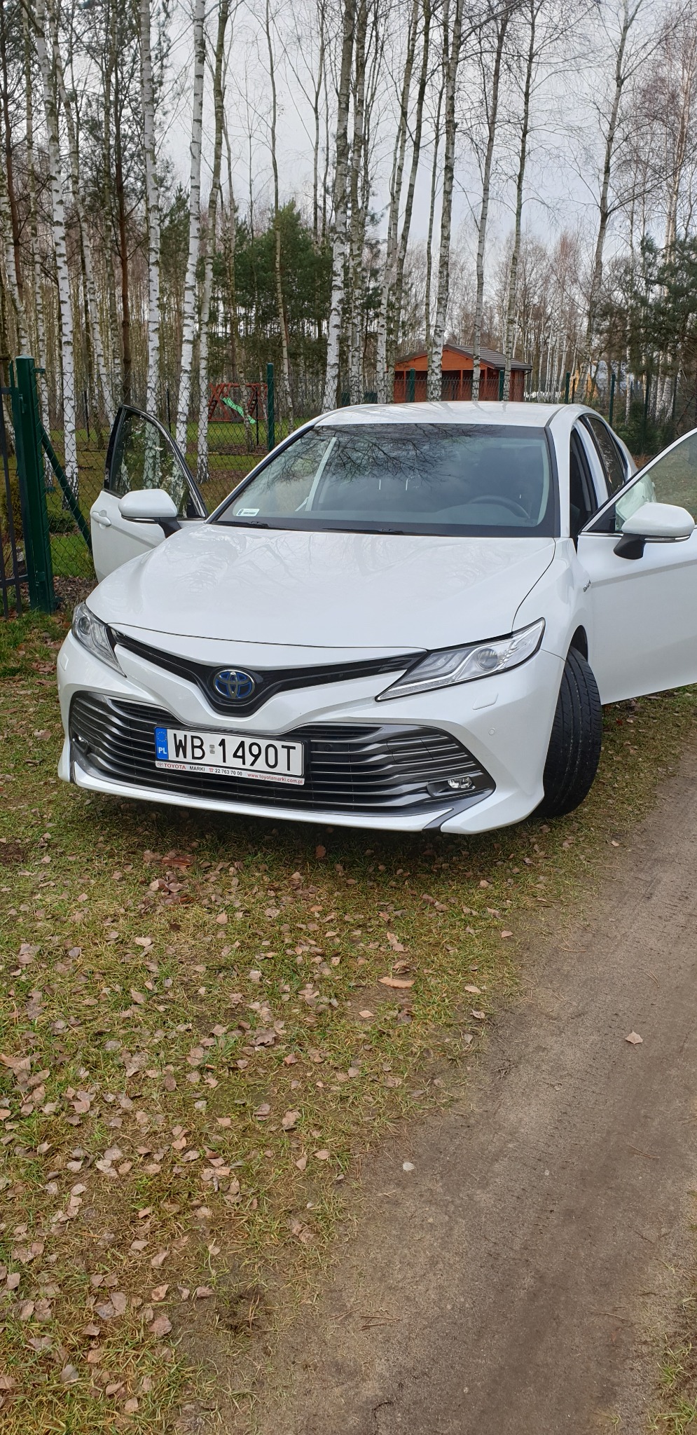Biała Toyota Camry z otwartymi drzwiami na tle brzozowego lasu. Widoczna tablica rejestracyjna WB 1490T. Auto stoi na trawie z opadłymi liśćmi.