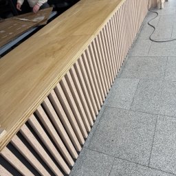 Woodsteel Pro Marcin Kamiński - Montaż blatu z lamelami na czarnym tle. Widoczny poziomica i pracownik w tle. Ujęcie z boku, ukazujące strukturę i detale wykończenia.