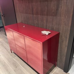 Woodsteel Pro Marcin Kamiński - Czerwona komoda z metalu na tle drewnianej ściany i półki. Minimalistyczny design, widoczne detale wykończenia, jasne oświetlenie. Nowoczesny styl.
