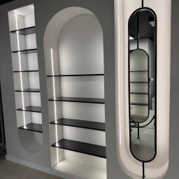 Woodsteel Pro Marcin Kamiński - Biała ściana z trzema wnękami: dwie z półkami podświetlonymi LED, trzecia z podświetlonym lustrem w czarnej ramie. Minimalistyczny design, nowoczesne oświetlenie.