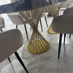 Woodsteel Pro Marcin Kamiński - Nowoczesny stół z czarnym blatem i złotą, spiralną podstawą, otoczony beżowymi krzesłami na tle marmurowej podłogi. Widoczne elementy dekoracyjne na stole.