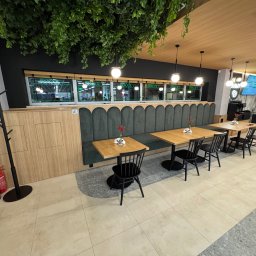 Woodsteel Pro Marcin Kamiński - Wnętrze kawiarni z drewnianymi stolikami, czarnymi krzesłami i zieloną tapicerowaną ławą pod oknami. Dekoracje roślinne na suficie i podłoga z jasnych płytek.