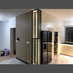 Nowoczesna kuchnia z zabudową: pionowe lamele na ścianie, przeszklona witryna z oświetleniem LED, błyszczące fronty szafek i marmurowa podłoga. Minimalistyczny design.