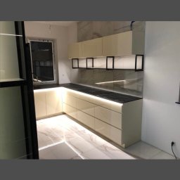 Beżowa kuchnia z podświetleniem LED, czarnym blatem i marmurową ścianą. Widok na okno i podłogę z płytek imitujących marmur. Nowoczesny design.