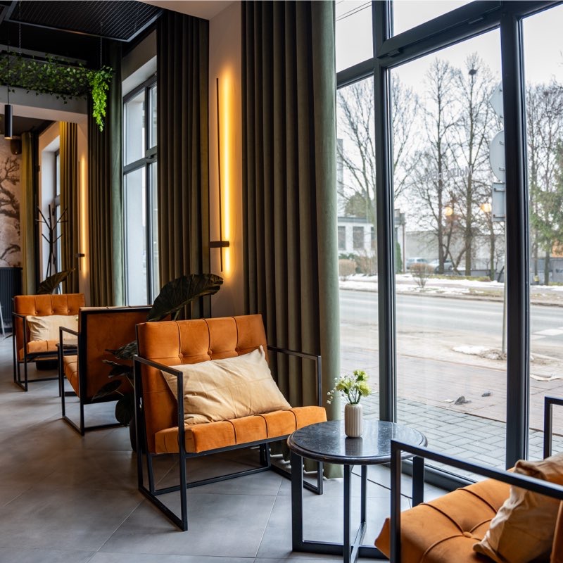 Pomarańczowe fotele z czarną ramą i beżową poduszką w nowoczesnym wnętrzu z dużymi oknami i zielonymi zasłonami. Na stoliku wazonik z kwiatami.