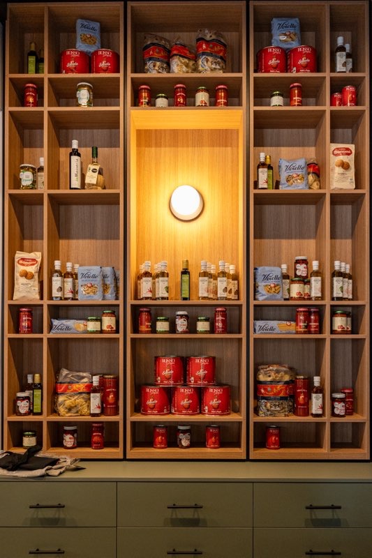 Drewniana zabudowa kuchenna z półkami, na których stoją produkty spożywcze w słoikach i puszkach. Centralna część z oświetleniem. Dolna część zabudowy z szufladami.