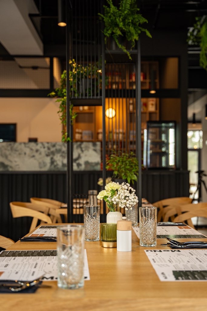 Stół w restauracji z dekoracjami: szklanki, wazoniki z kwiatami, menu. W tle regał z roślinami i oświetleniem, tworzący elegancki wystrój wnętrza.