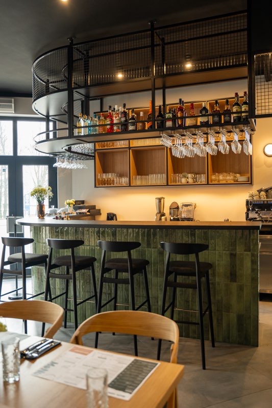 Zaokrąglony bar z zielonymi płytkami, czarnymi hokerami i metalową konstrukcją nad blatem. Na półkach butelki i szklanki, w tle okno i stoliki.