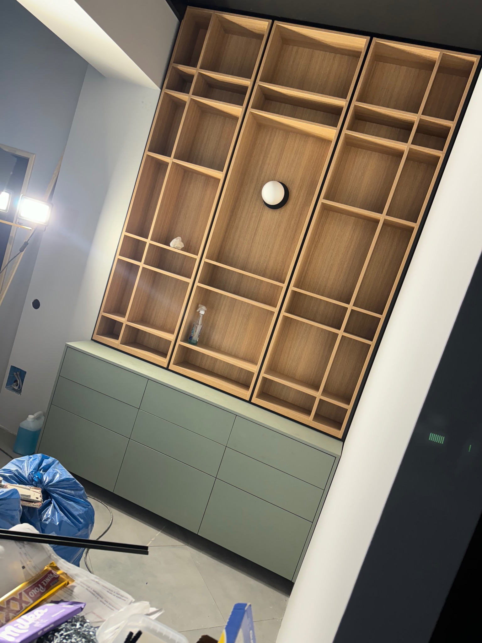 Wysoka zabudowa z drewnianymi półkami i zielonymi szafkami na dole, w trakcie montażu. Widoczne narzędzia i materiały budowlane. Nietypowy kąt ujęcia.