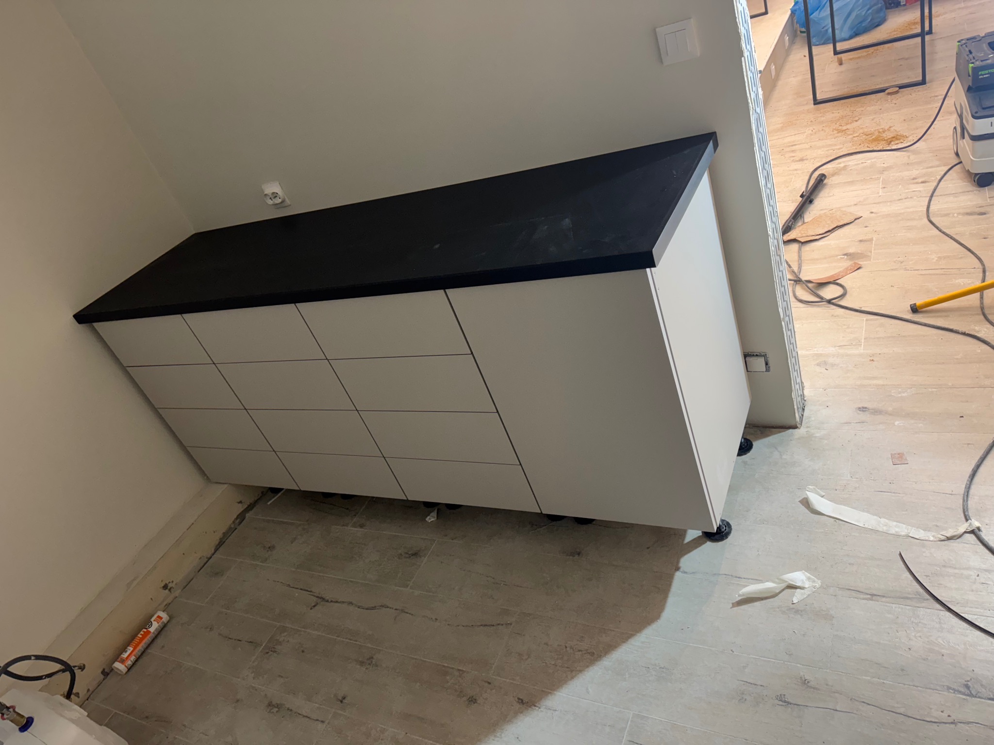 Biała szafka z czarnym blatem na kółkach, ustawiona przy ścianie w trakcie wykańczania wnętrza. Widoczne narzędzia i resztki po pracy.