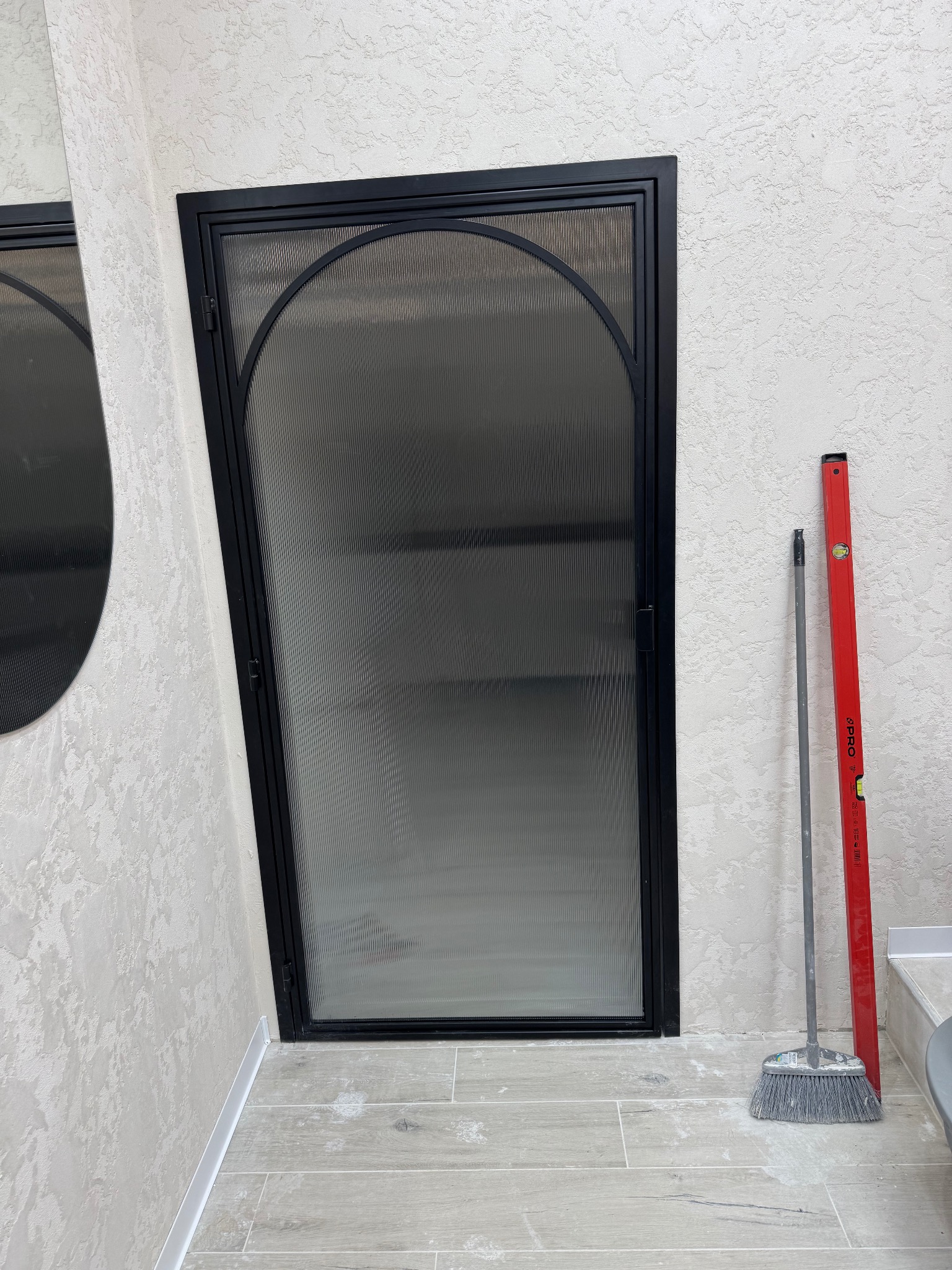 Nowoczesne drzwi z czarną ramą i ryflowanym szkłem, łukowate u góry, stojące w wykończonym wnętrzu. Obok poziomica i miotła.