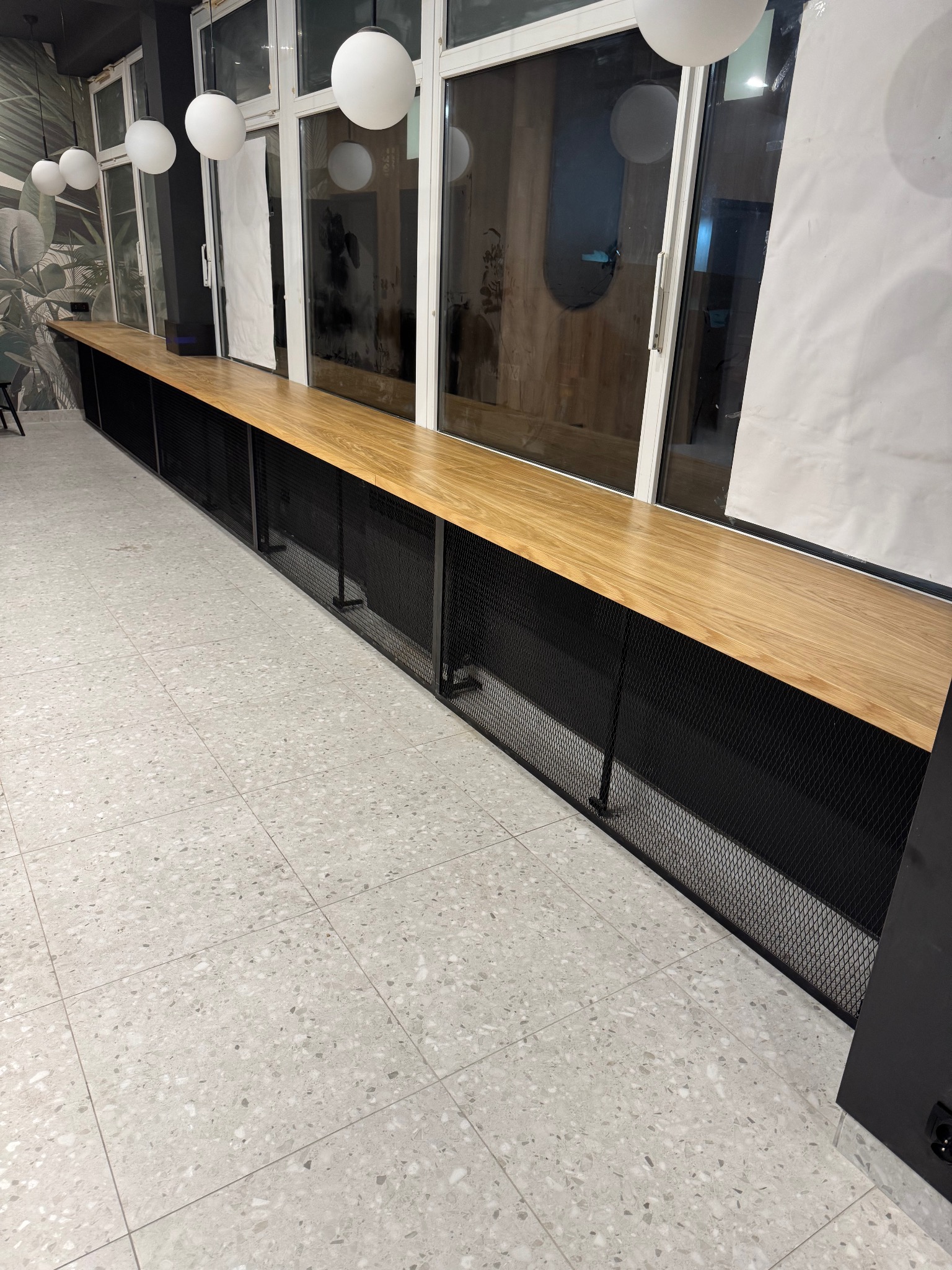 Długa, drewniana lada z czarną, metalową siatką u podstawy, umieszczona wzdłuż okien. Białe, kuliste lampy wiszą z sufitu. Jasna, wzorzysta podłoga.