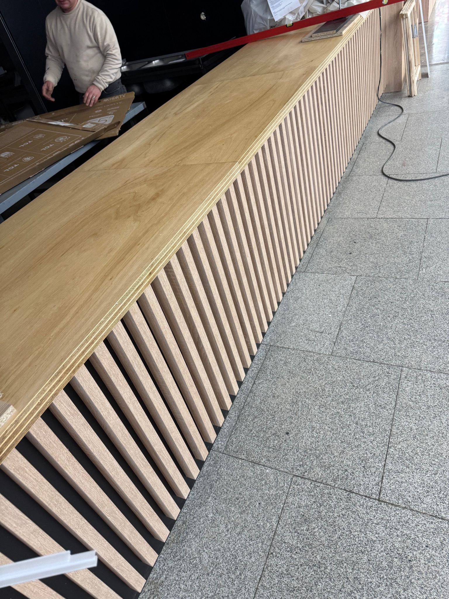 Montaż blatu z lamelami na czarnym tle. Widoczny poziomica i pracownik w tle. Ujęcie z boku, ukazujące strukturę i detale wykończenia.