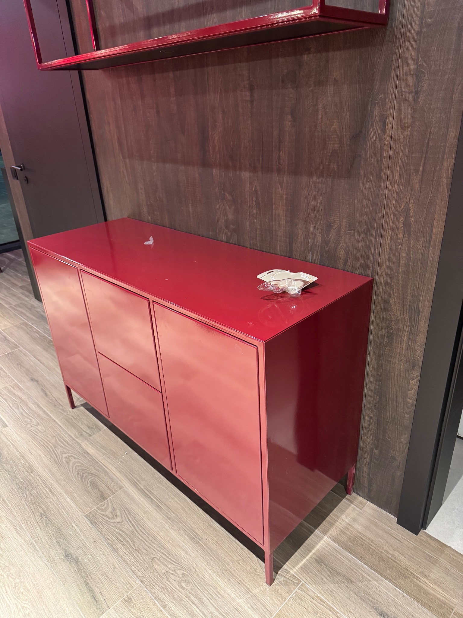 Czerwona komoda z metalu na tle drewnianej ściany i półki. Minimalistyczny design, widoczne detale wykończenia, jasne oświetlenie. Nowoczesny styl.