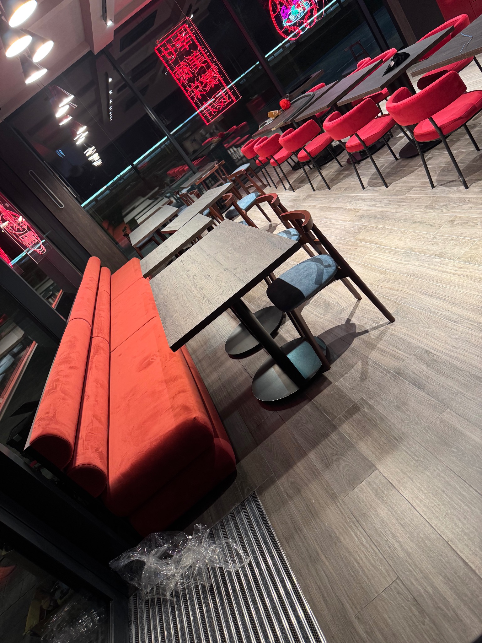 Wnętrze restauracji z rzędami stołów i krzeseł. Czerwona, długa sofa wzdłuż ściany. Neonowe dekoracje na ścianach. Podłoga wyłożona panelami imitującymi drewno.