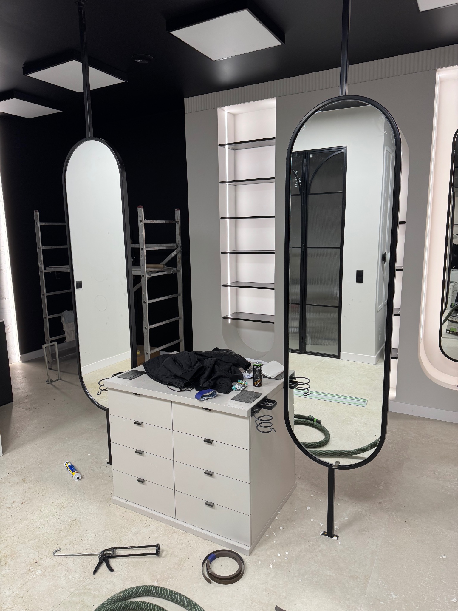 Montaż wiszących luster w czarnej ramie w salonie. Widoczne narzędzia, komoda i drabina. W tle półki i czarna ściana. Prace wykończeniowe w toku.