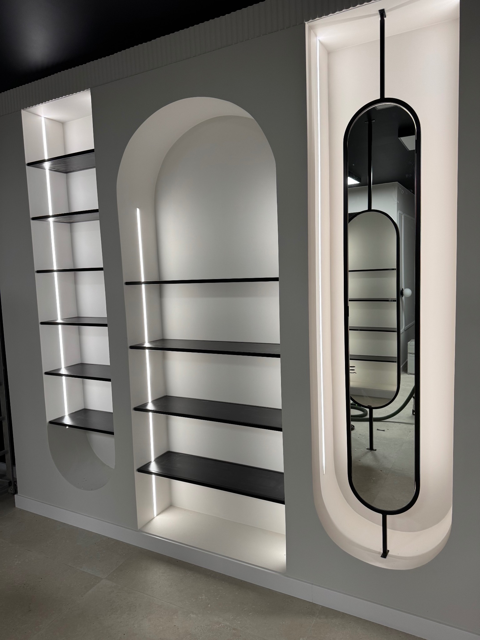 Biała ściana z trzema wnękami: dwie z półkami podświetlonymi LED, trzecia z podświetlonym lustrem w czarnej ramie. Minimalistyczny design, nowoczesne oświetlenie.