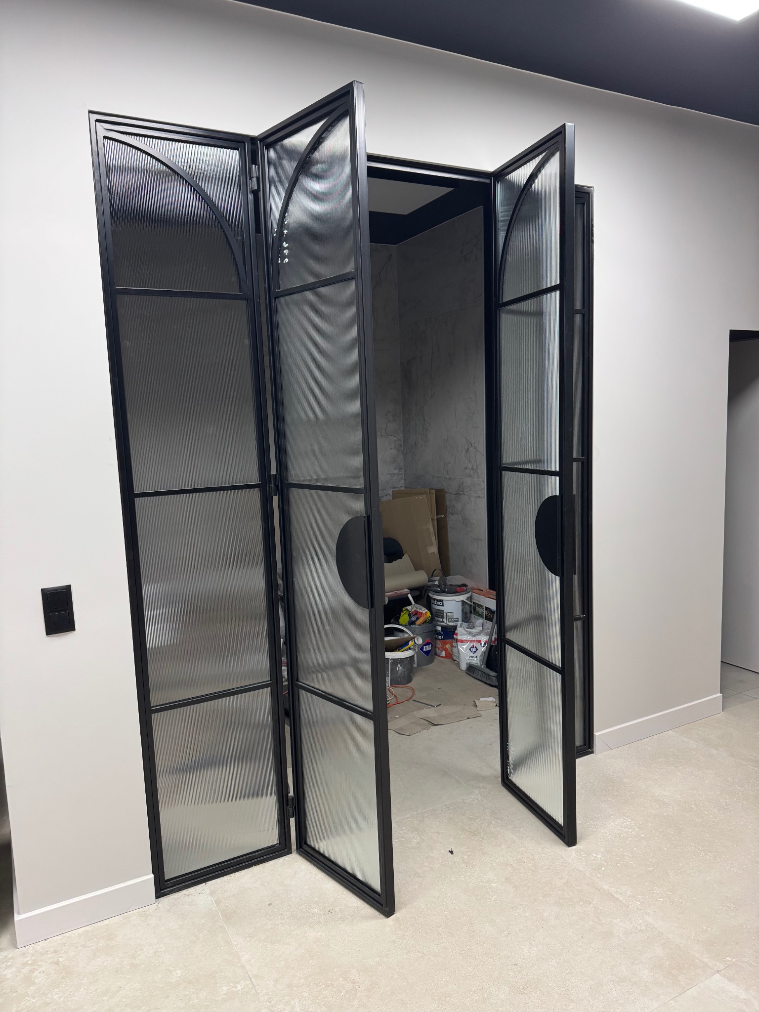 Nowoczesne, składane drzwi loftowe z czarną ramą i ryflowanym szkłem, otwarte do wnętrza w trakcie wykańczania. Widoczne puszki z farbą i narzędzia.