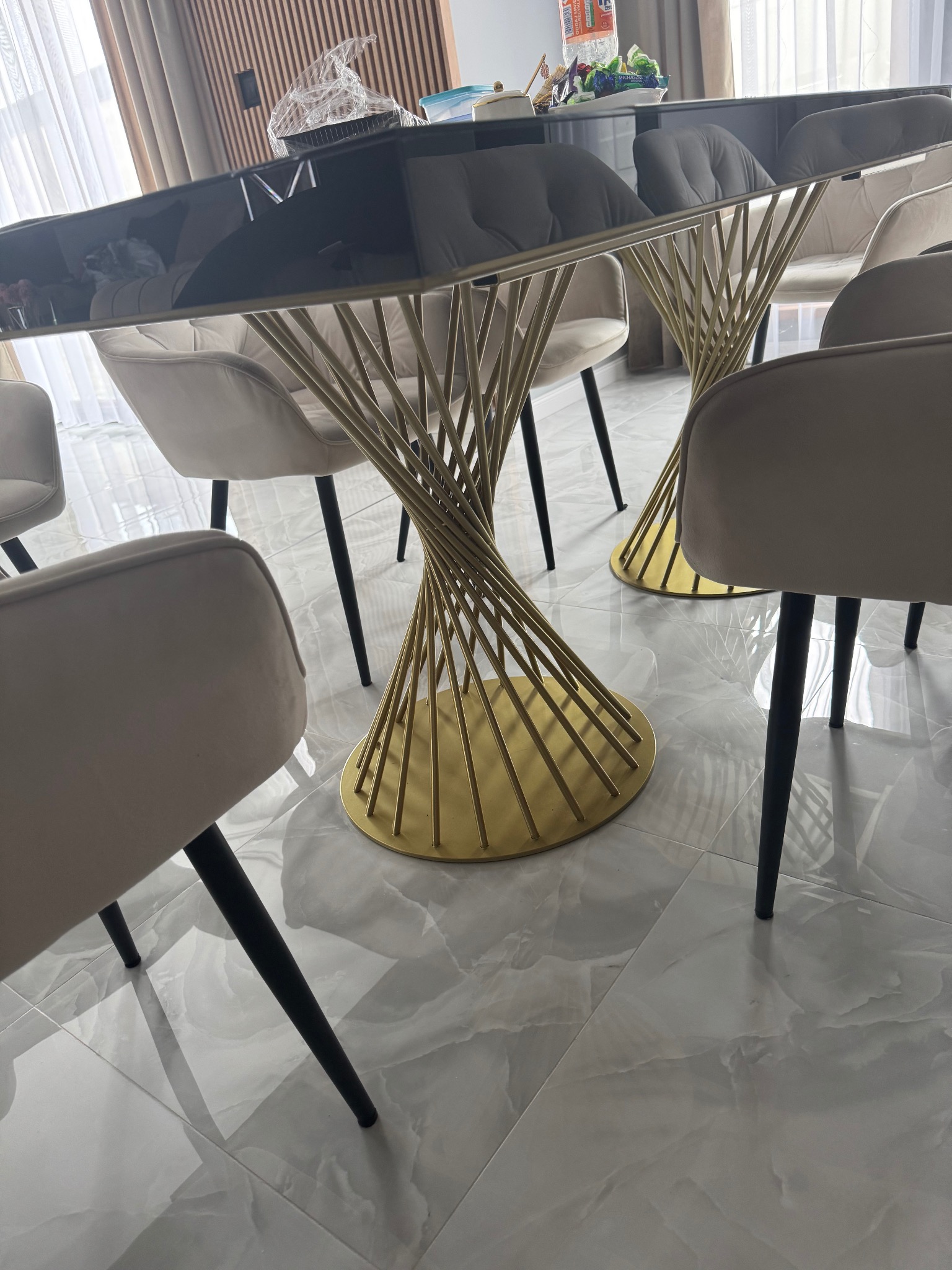 Nowoczesny stół z czarnym blatem i złotą, spiralną podstawą, otoczony beżowymi krzesłami na tle marmurowej podłogi. Widoczne elementy dekoracyjne na stole.