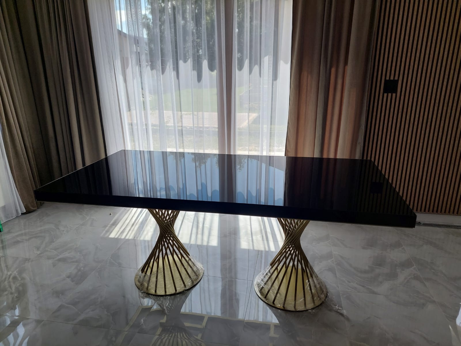 Czarny stół z połyskiem i złotymi, spiralnymi nogami na tle okna z zasłonami i drewnianej ściany. Nowoczesny design, eleganckie wykończenie, widoczne odbicia światła.