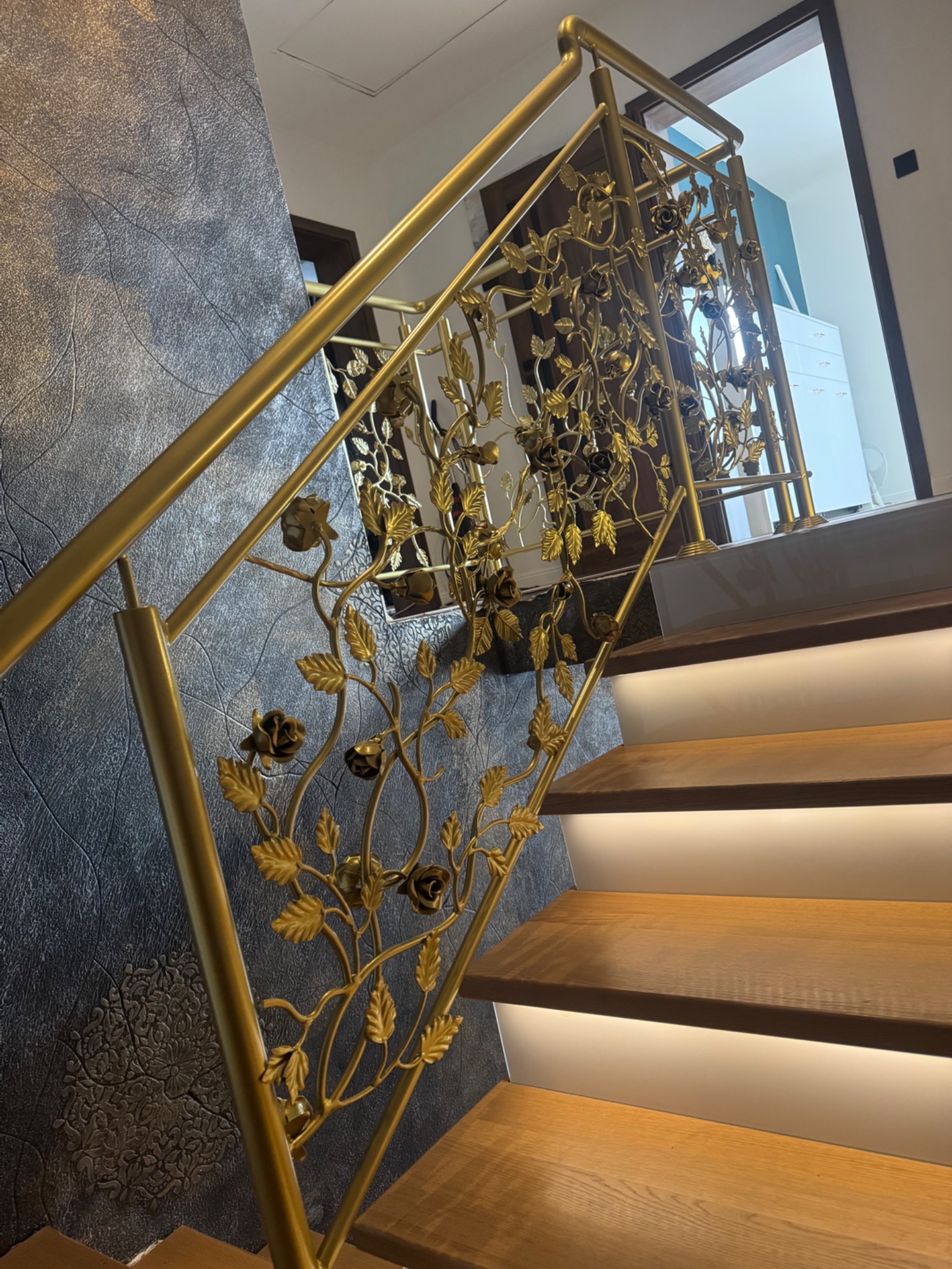 Złota, zdobiona balustrada schodowa z motywem roślinnym, oświetlone stopnie drewniane, ciemna ściana z fakturą. Detal wykonania i elegancja.