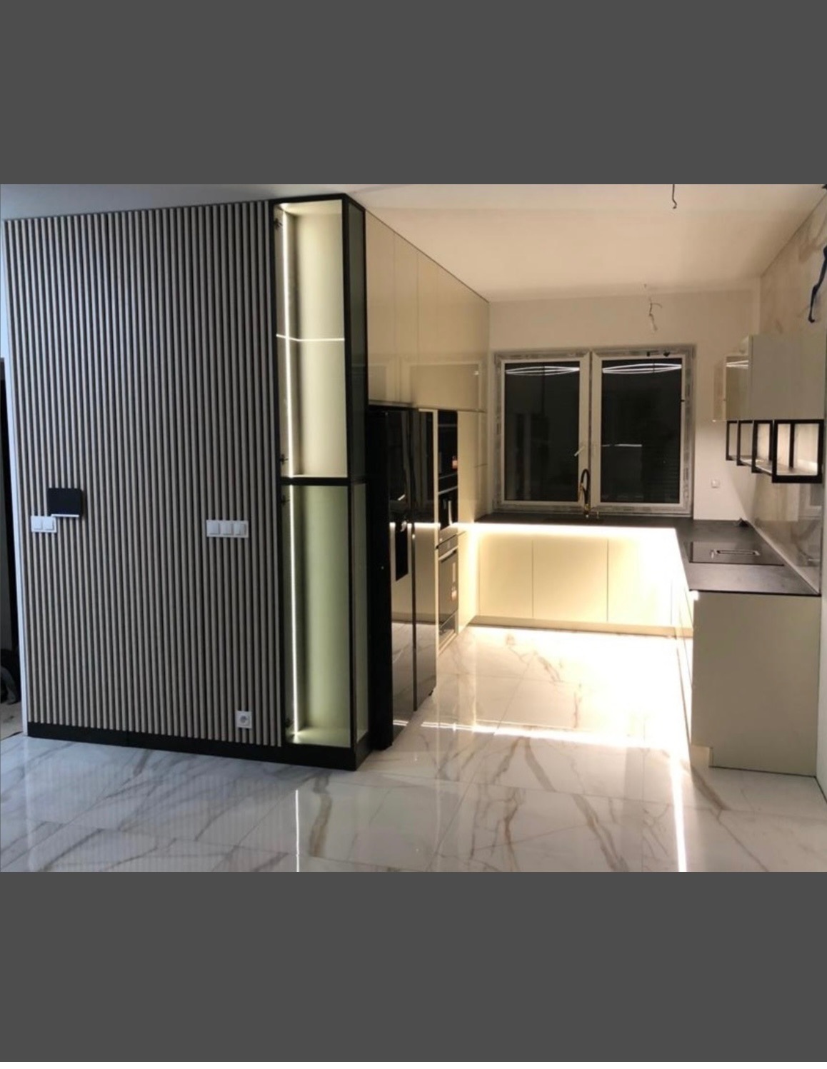 Nowoczesna kuchnia z białymi frontami, podświetleniem LED i marmurową podłogą. Na ścianie dekoracyjne lamele w odcieniach szarości. Minimalistyczny design.