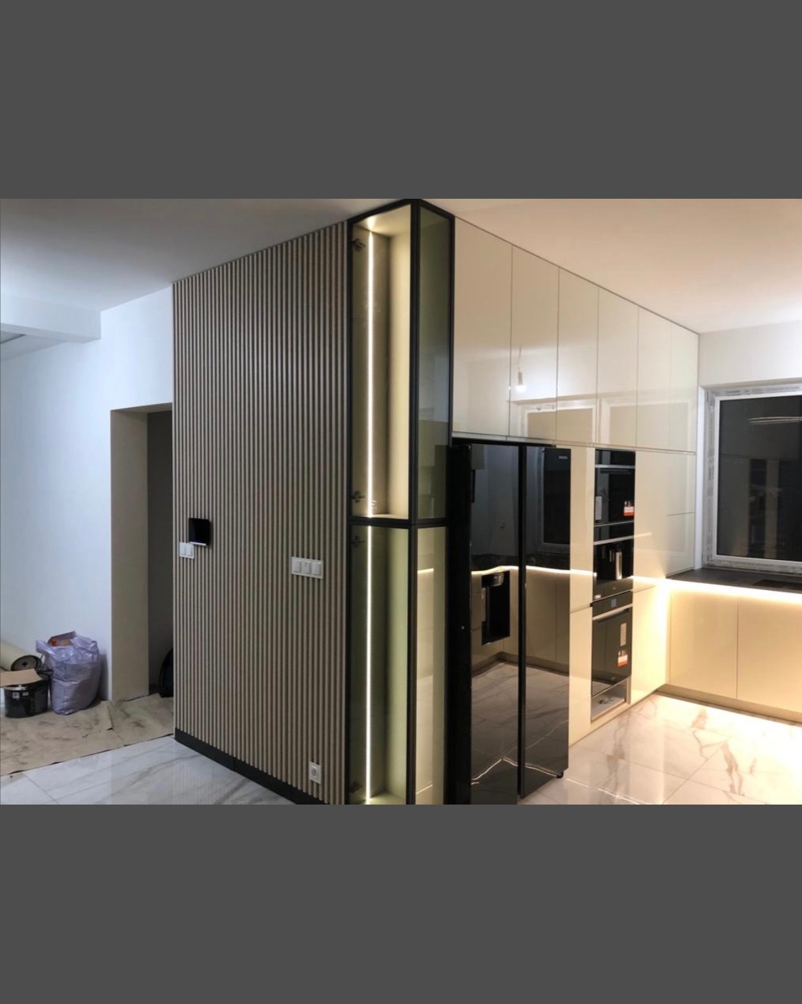 Nowoczesna kuchnia z zabudową: pionowe lamele na ścianie, przeszklona witryna z oświetleniem LED, błyszczące fronty szafek i marmurowa podłoga. Minimalistyczny design.