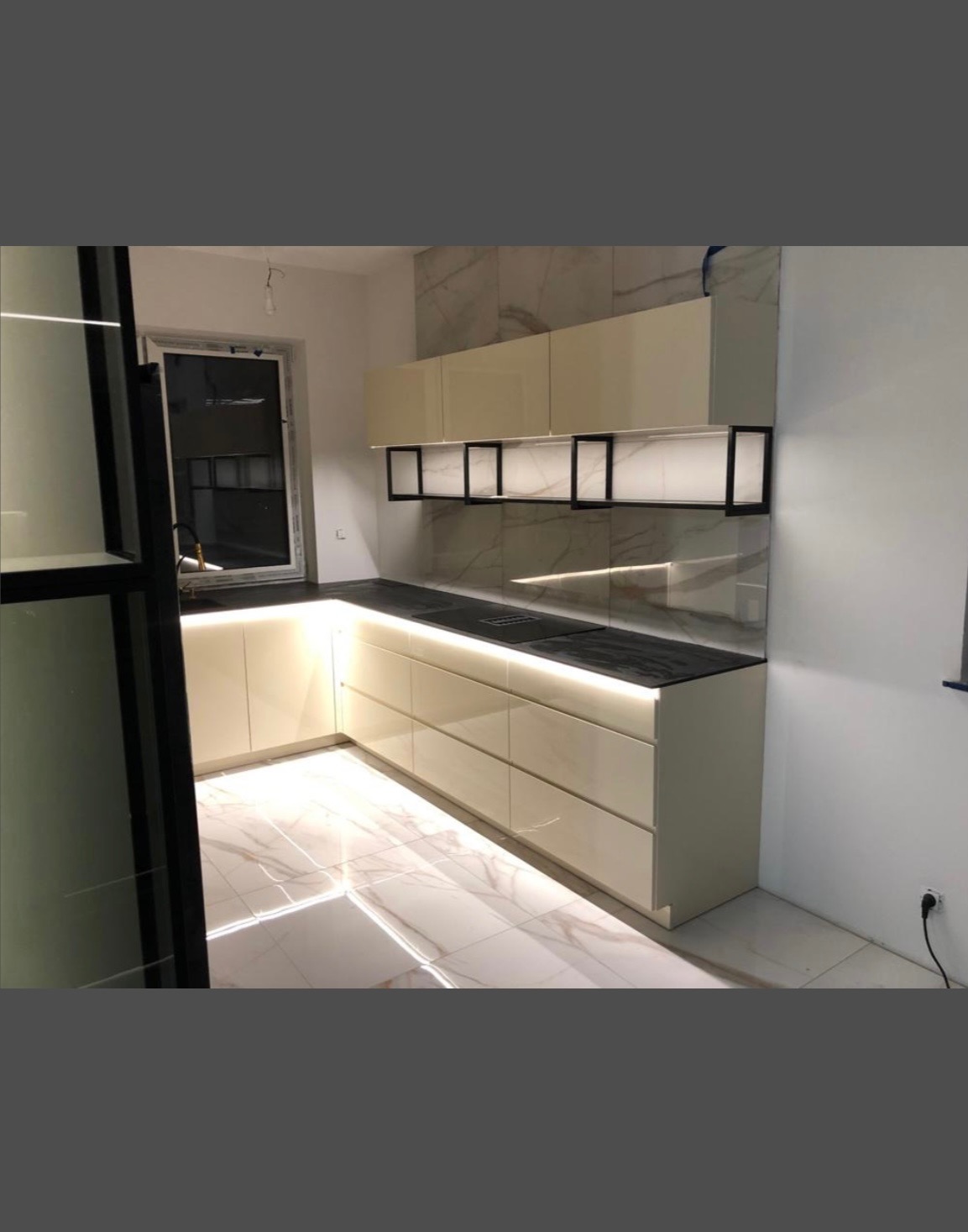 Beżowa kuchnia z podświetleniem LED, czarnym blatem i marmurową ścianą. Widok na okno i podłogę z płytek imitujących marmur. Nowoczesny design.