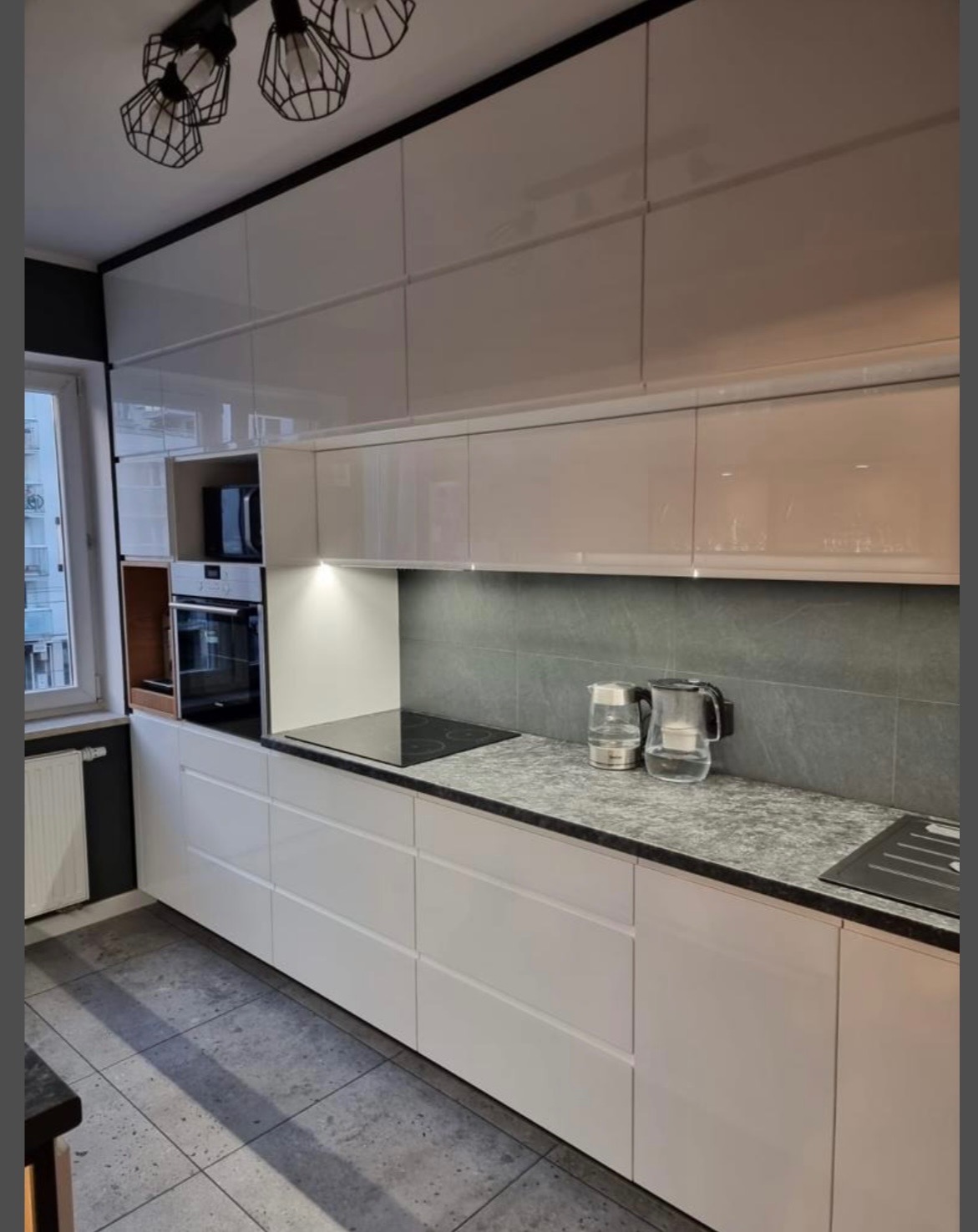 Biała, nowoczesna kuchnia z błyszczącymi frontami, szarym blatem i płytkami. Widoczne dwa czajniki elektryczne i oświetlenie podszafkowe.