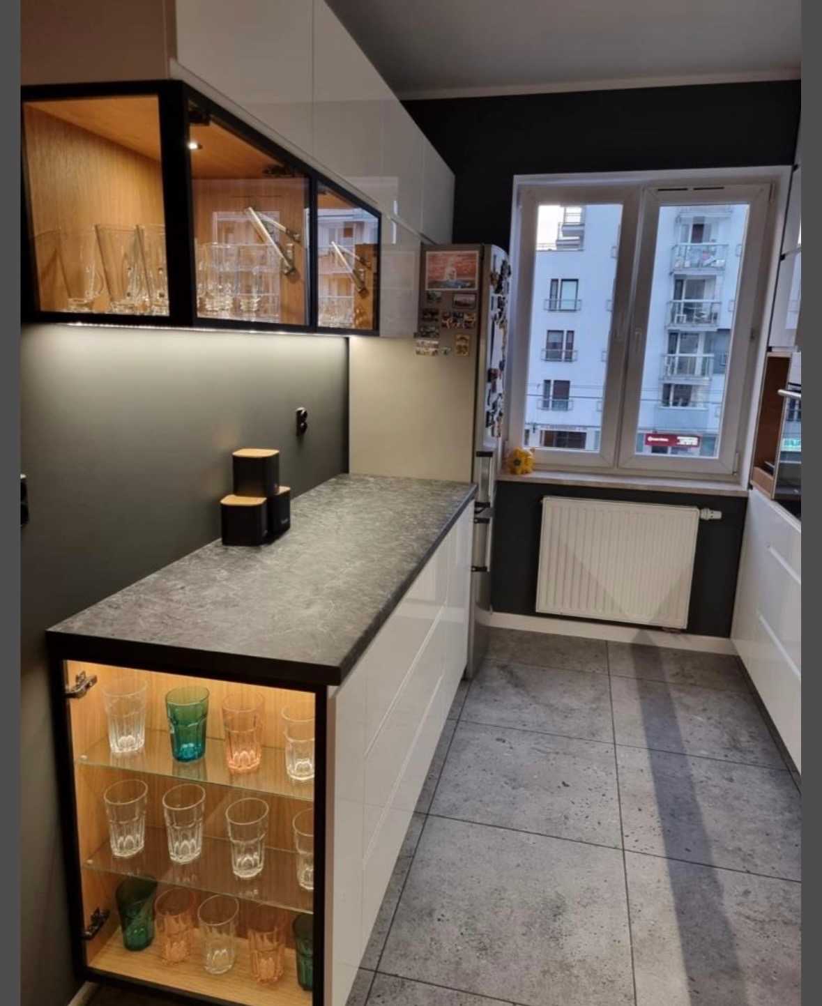 Nowoczesna kuchnia z szarym blatem i białymi szafkami. Podświetlane półki z szkła eksponują kolekcję szklanek. Widok na budynek przez okno.