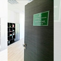 Kancelaria Adwokacka Adwokat Martynian Jasek - Otwarta, ciemna drewniana drzwi do kancelarii adwokackiej z zieloną tabliczką informacyjną z nazwiskiem adwokata Martyniaka Jaska i numerem telefonu. W tle regał z dokumentami.