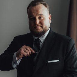 Kancelaria Adwokacka Adwokat Martynian Jasek - Portret mężczyzny w czarnym garniturze, poprawiającego spinkę do krawata. Stonowane tło, profesjonalny wygląd, subtelne oświetlenie.