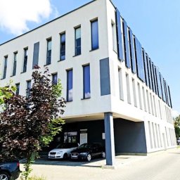 Adwokat Martynian Jasek Kancelaria Adwokacka Lublin - Nowoczesny biały budynek biurowy z pionowymi panelami fotowoltaicznymi na bocznej elewacji, dużymi oknami i zadaszonym parkingiem z dwoma samochodami, otoczony zielenią.