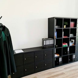 Adwokat Martynian Jasek Kancelaria Adwokacka Lublin - Czarna toga prawnicza z zielonym żabotem wisi na wieszaku, obok nowoczesne, czarne regały modułowe wypełnione segregatorami i książkami, na tle białej ściany i jasnej podłogi.