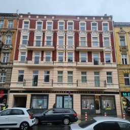 PATRYK PEJSKI - Odnowiona kamienica z balkonami i dekoracyjną elewacją w odcieniach czerwieni i beżu, z widocznym szyldem firmy na parterze. Ulica z zaparkowanymi autami.