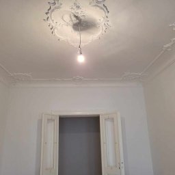 PATRYK PEJSKI - Białe, odnowione wnętrze z dekoracyjnymi sztukateriami na suficie i ścianach, oświetlone żarówką wiszącą. Otwarty widok na sąsiednie pomieszczenie.