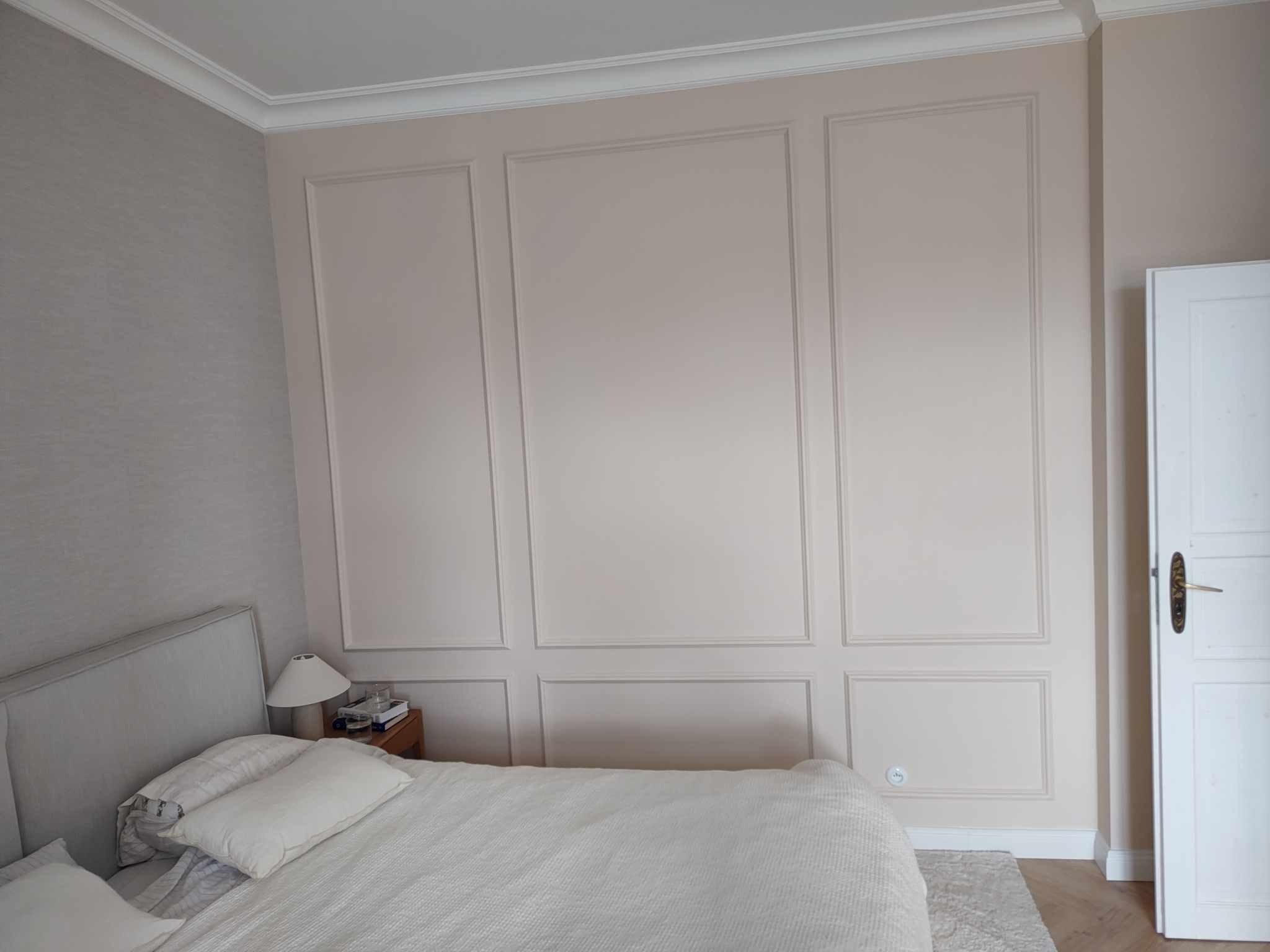 Elegancka ściana z dekoracyjnymi panelami w odcieniach beżu i bieli, widoczna w sypialni z łóżkiem i lampką nocną. Klasyczny wystrój wnętrza.