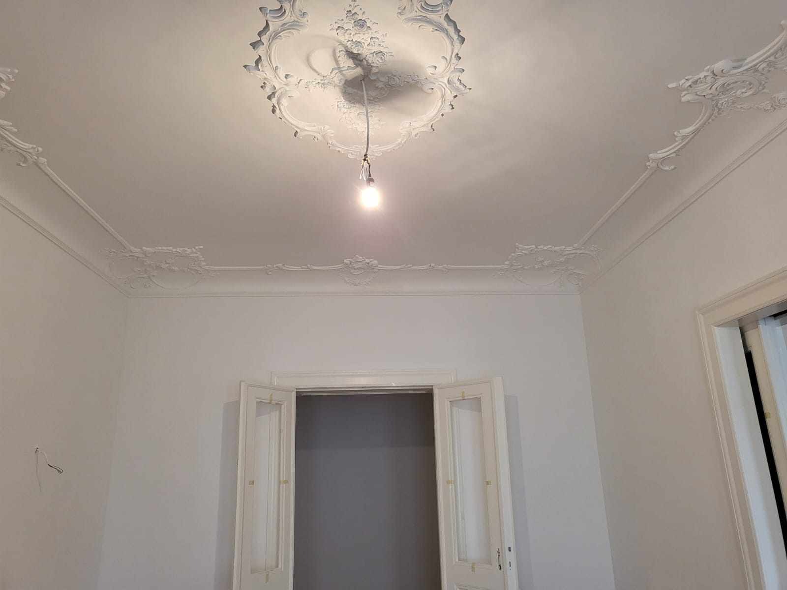 Białe, odnowione wnętrze z dekoracyjnymi sztukateriami na suficie i ścianach, oświetlone żarówką wiszącą. Otwarty widok na sąsiednie pomieszczenie.